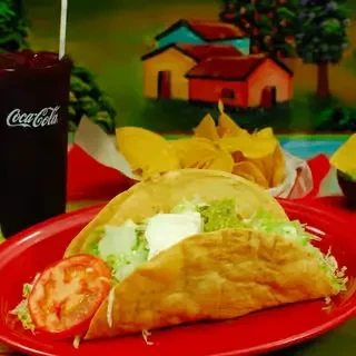 Fried Mexican tacos, soda, chips, against a colorful mural.