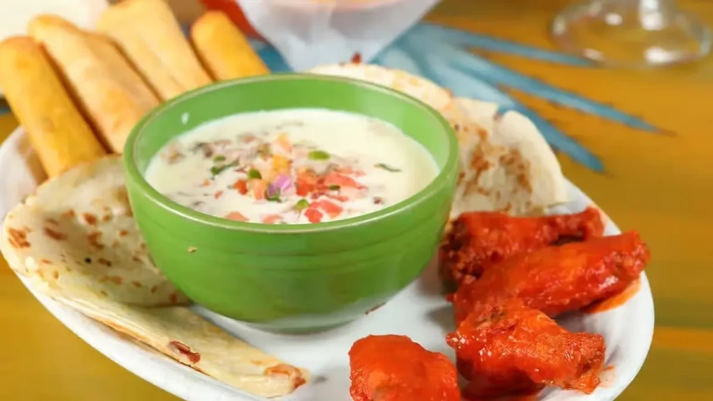White plate with green dip, golden breadsticks, flatbreads, and red chicken wings.