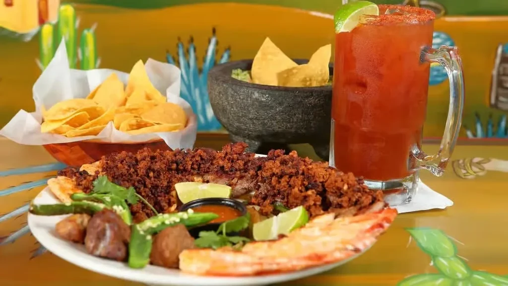 Mexican restaurant table with meat dish, guacamole, chips, and michelada.