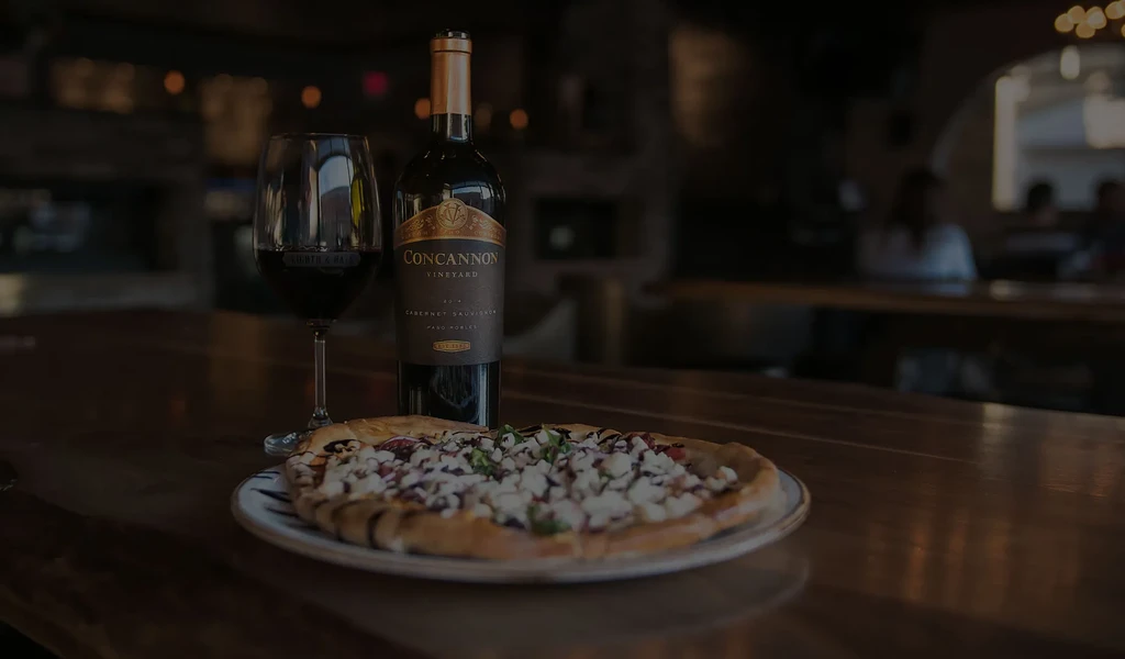 Rustic pizza, red wine, and wine bottle in a dimly lit restaurant.