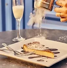 Hand dusts powdered sugar over chocolate-drizzled cheesecake slice.