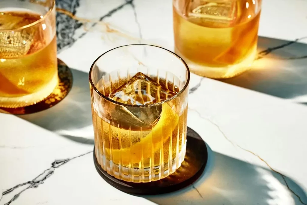 Three amber cocktails with ice and lemon twists on white marble.