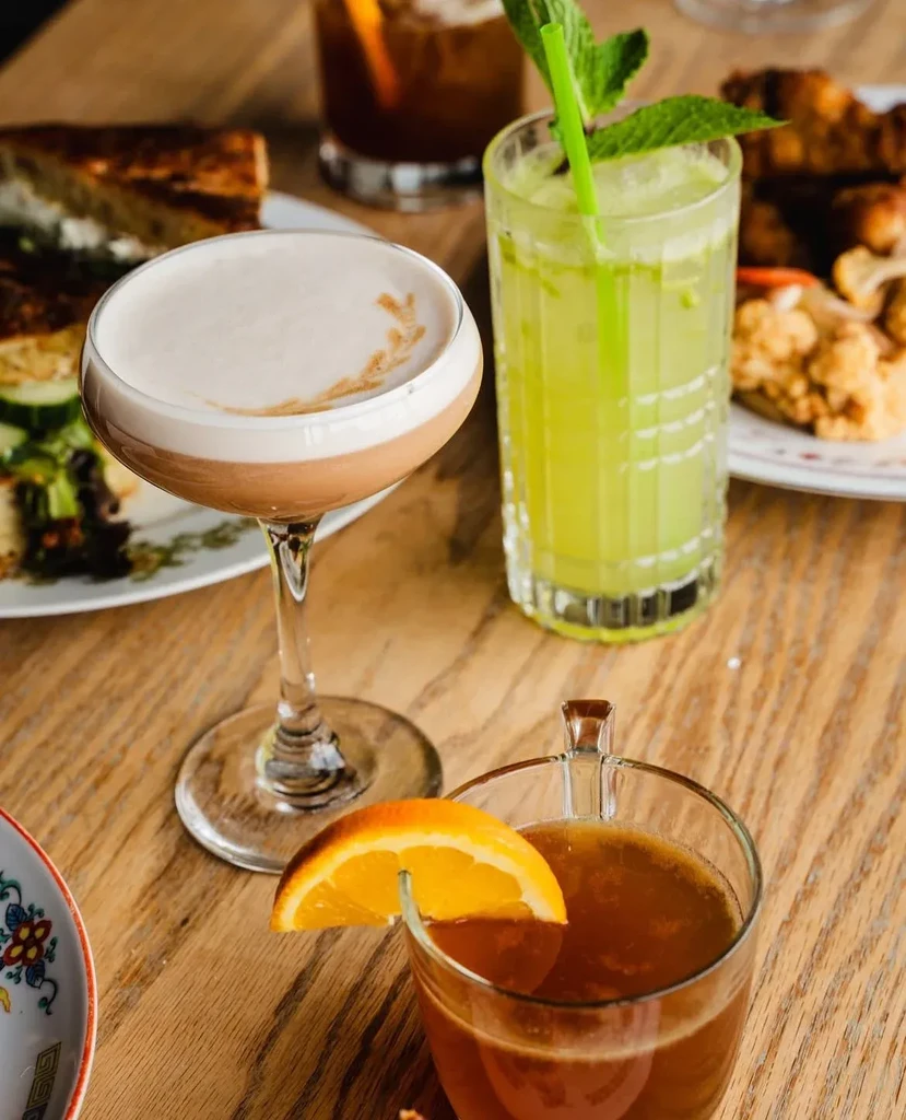 Three artfully presented drinks and food on a light wooden table.
