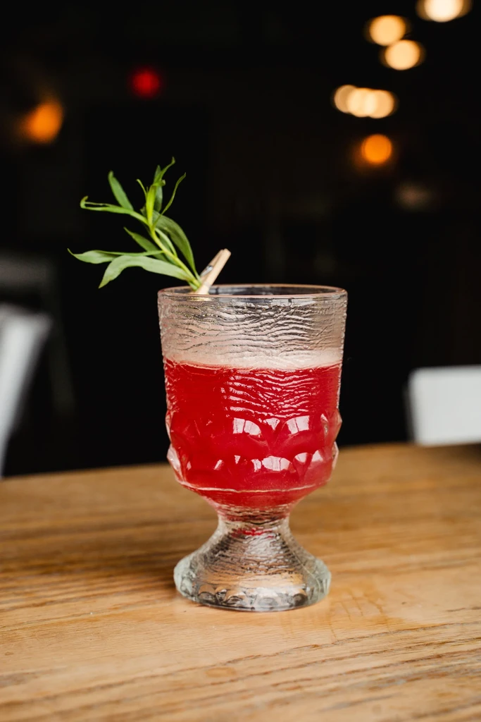 Red cocktail in textured glass with herb sprig on wooden table.