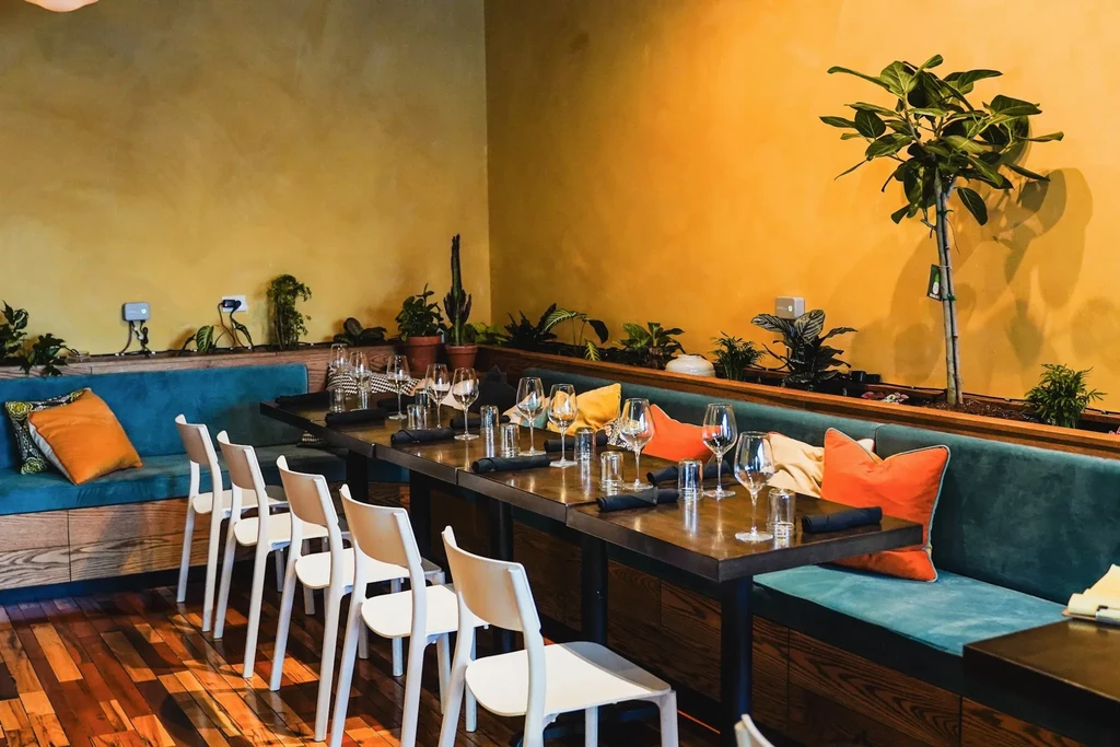 Restaurant interior with golden walls, teal banquette, plants, and set tables.
