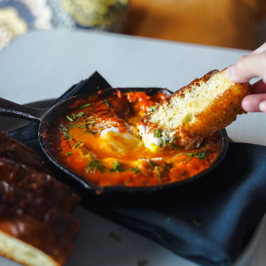 Hand dips toast into eggs in cast-iron skillet with tomato sauce.