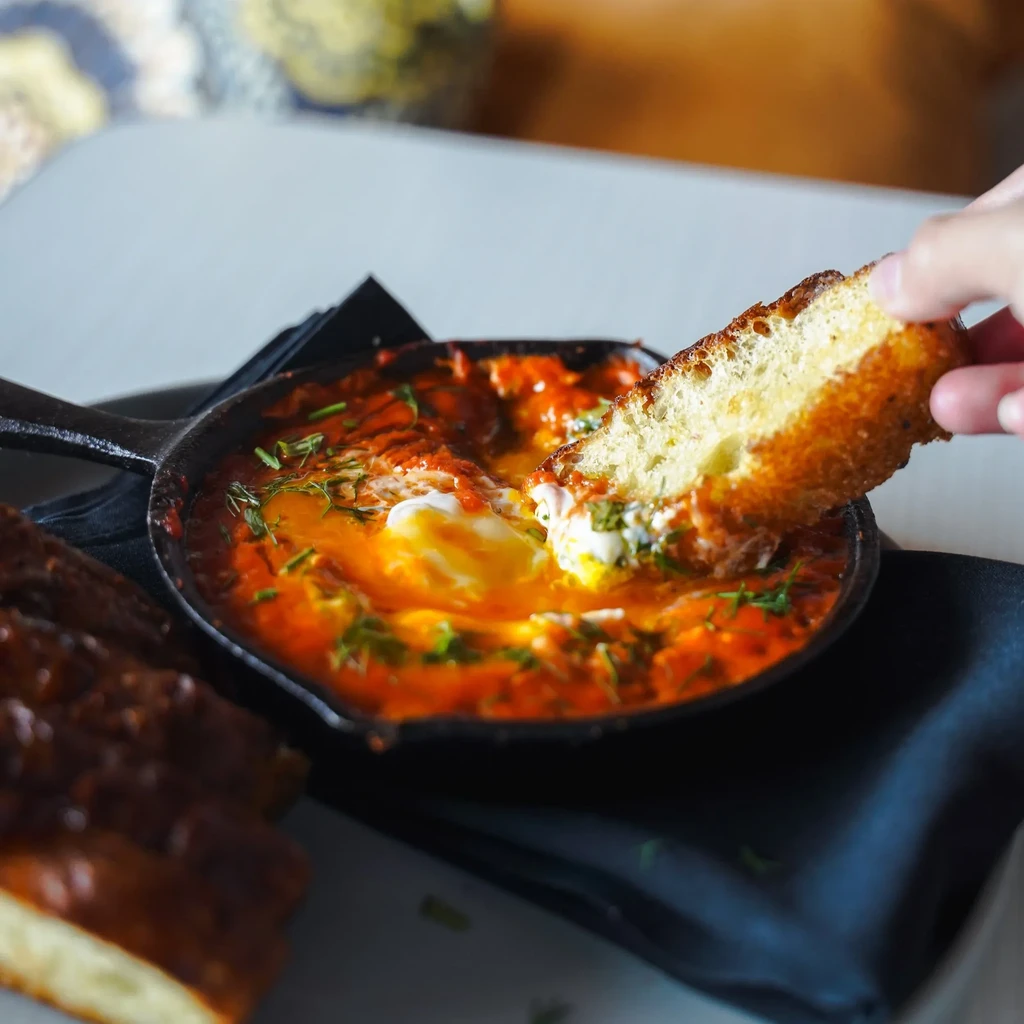Hand dipping bread into a skillet of eggs and tomato sauce.