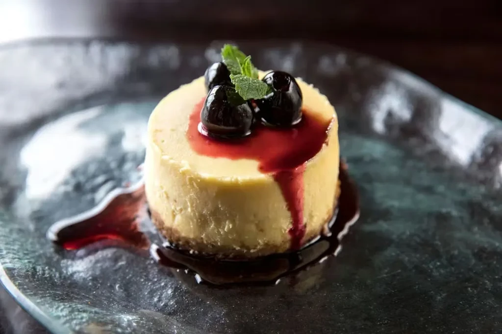 Round cheesecake with red berry sauce, dark berries, and mint.