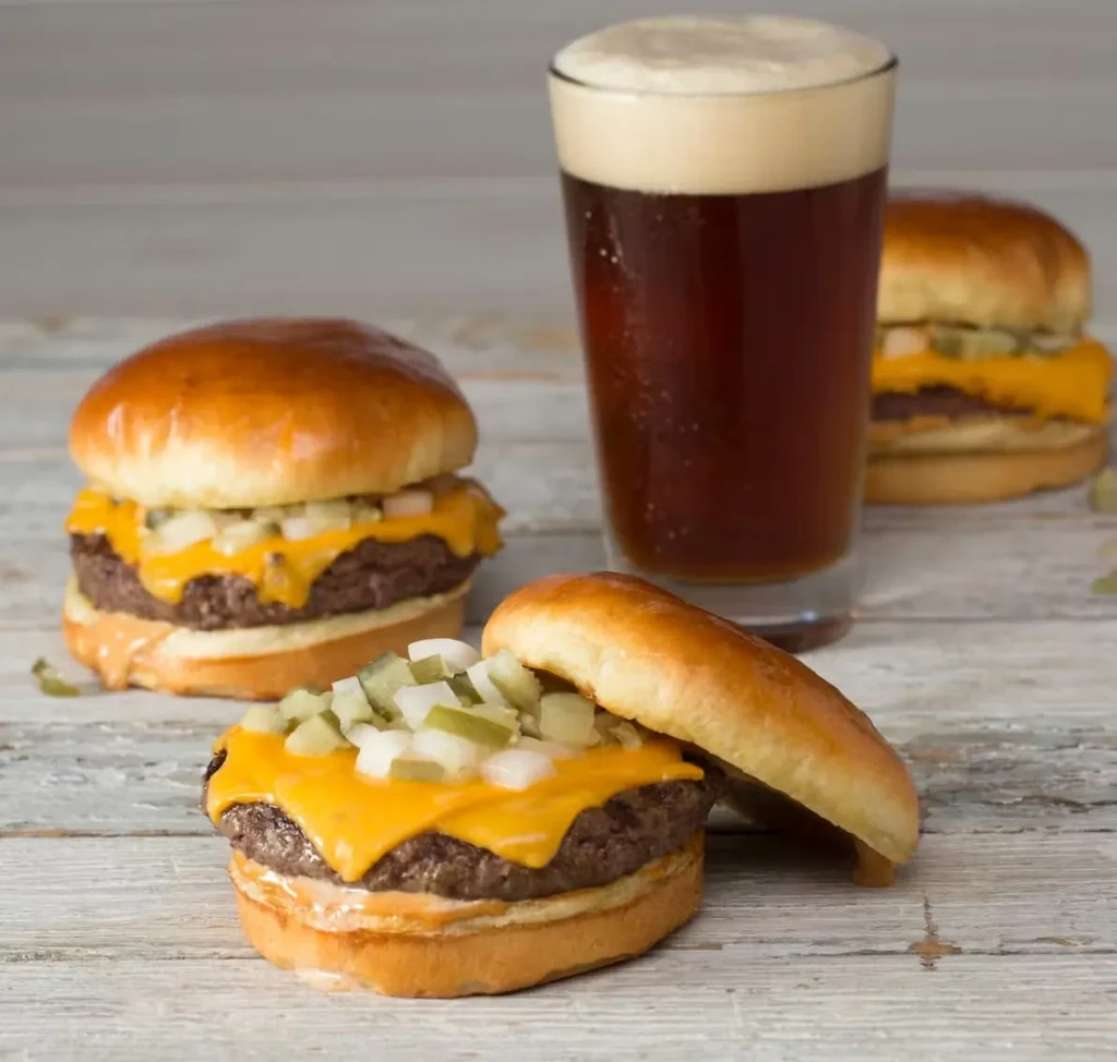 Three mini cheeseburgers and a pint of dark beer on wood.