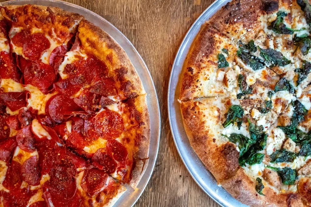Two distinct pizzas: pepperoni, and white sauce chicken spinach, on wood.