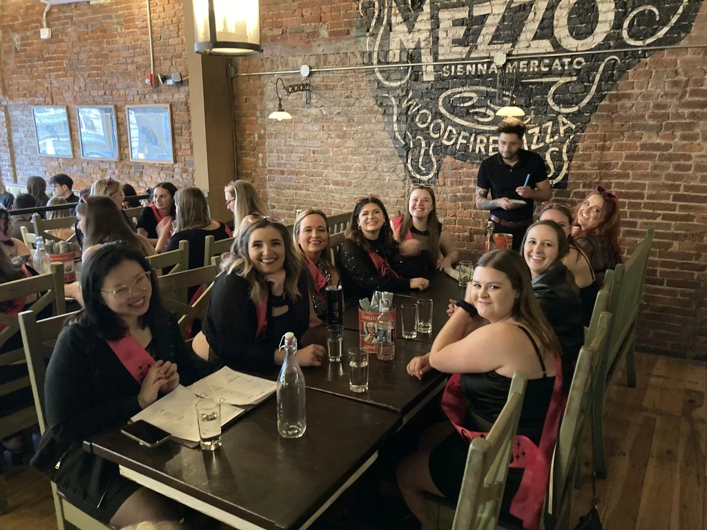 Women in pink sashes smiling in a rustic pizza restaurant.