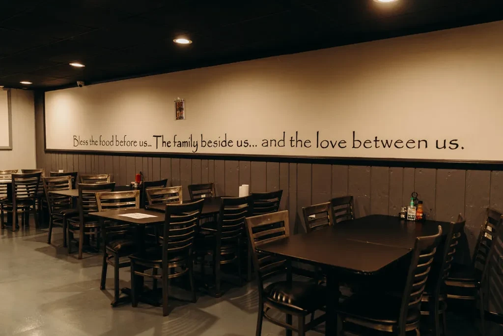 Empty restaurant dining room with dark wood tables, chairs, and wall quote.