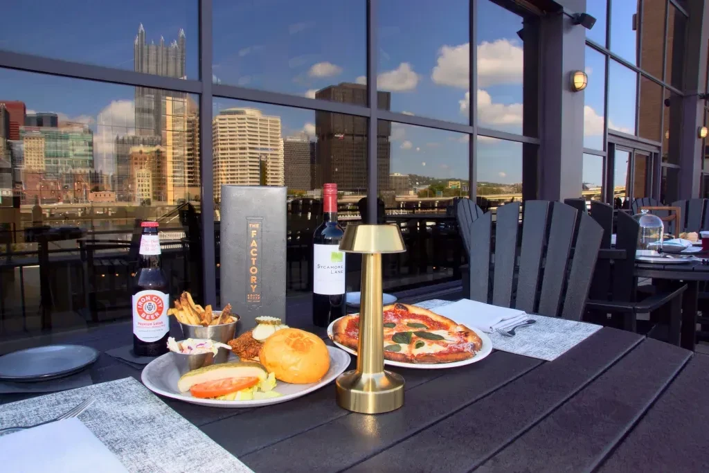 Outdoor dining table with chicken, pizza, beer, and Pittsburgh skyline reflected.