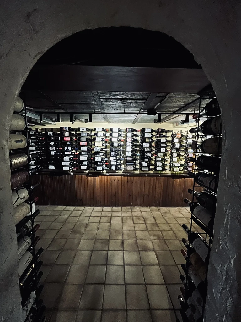 Rustic wine cellar with bottle-filled walls, seen through an archway.