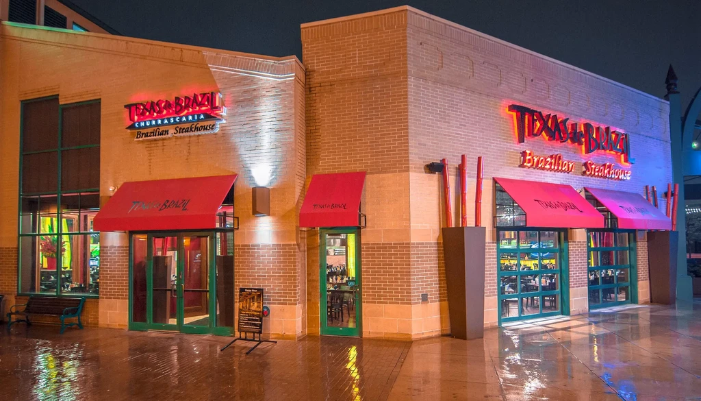 Exterior of Texas de Brazil steakhouse at night, neon signs, wet ground.