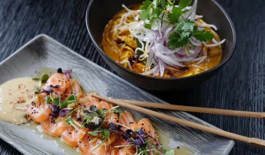 Top-down view of salmon sashimi and noodle soup on a dark table.