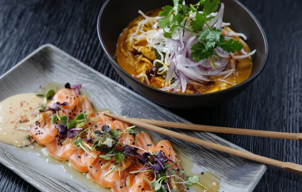 Plated salmon sashimi and a richly garnished noodle dish on dark surface.