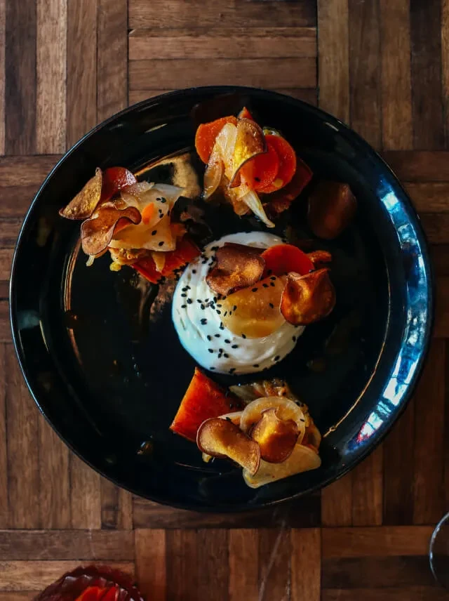Artfully plated dish with creamy spread, black seeds, and crispy root chips.