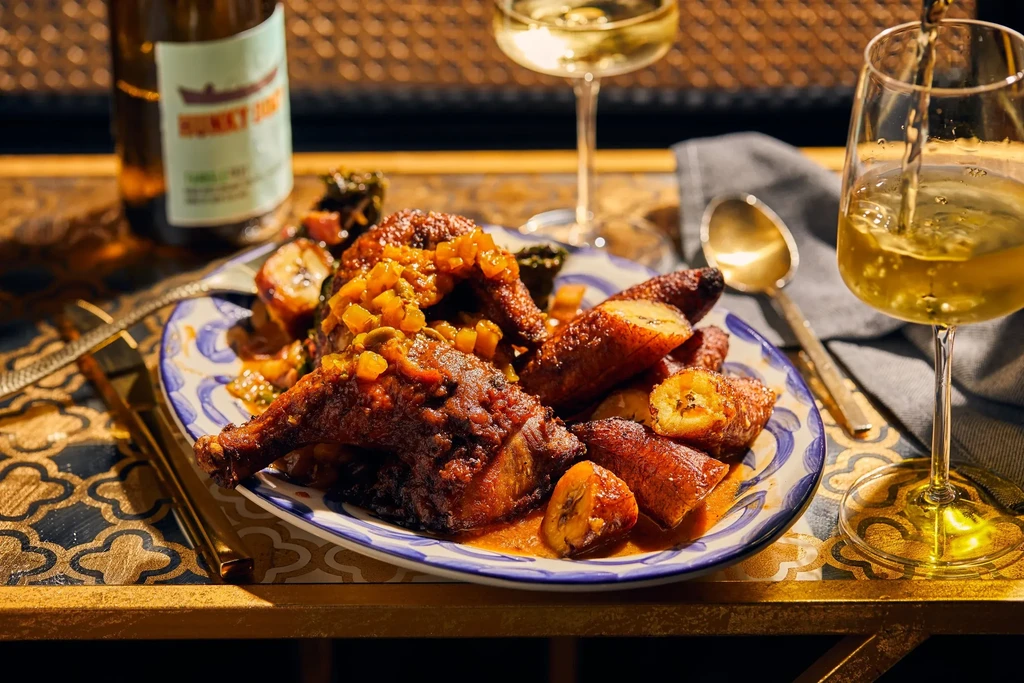 Roasted poultry and plantains with sauce on a patterned plate, with pouring wine.