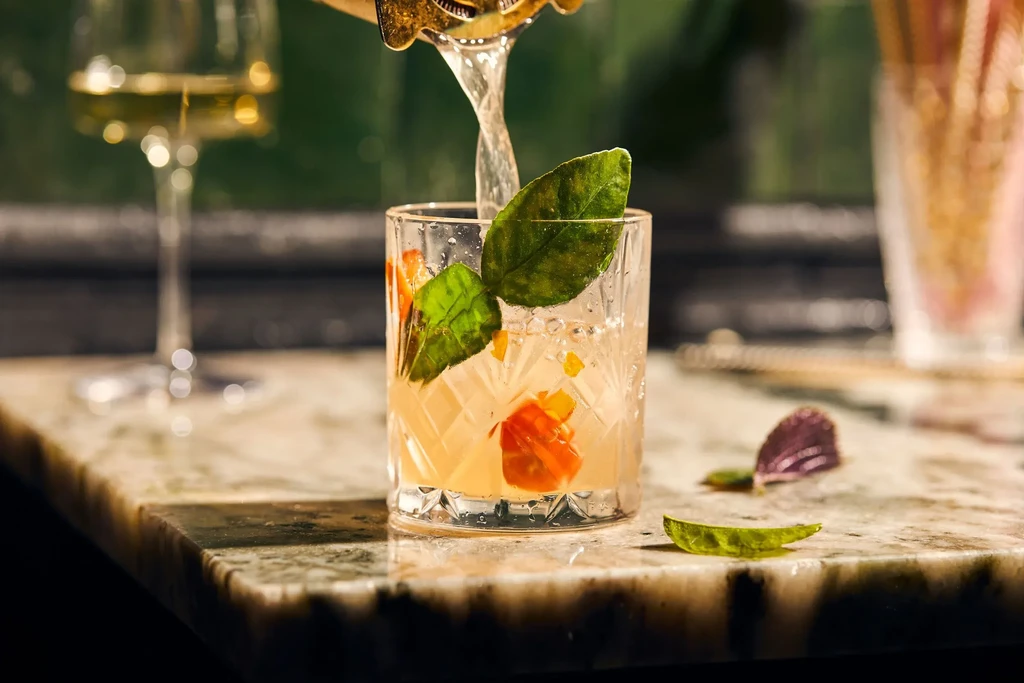 Cocktail with green leaves and fruit being poured into textured glass.