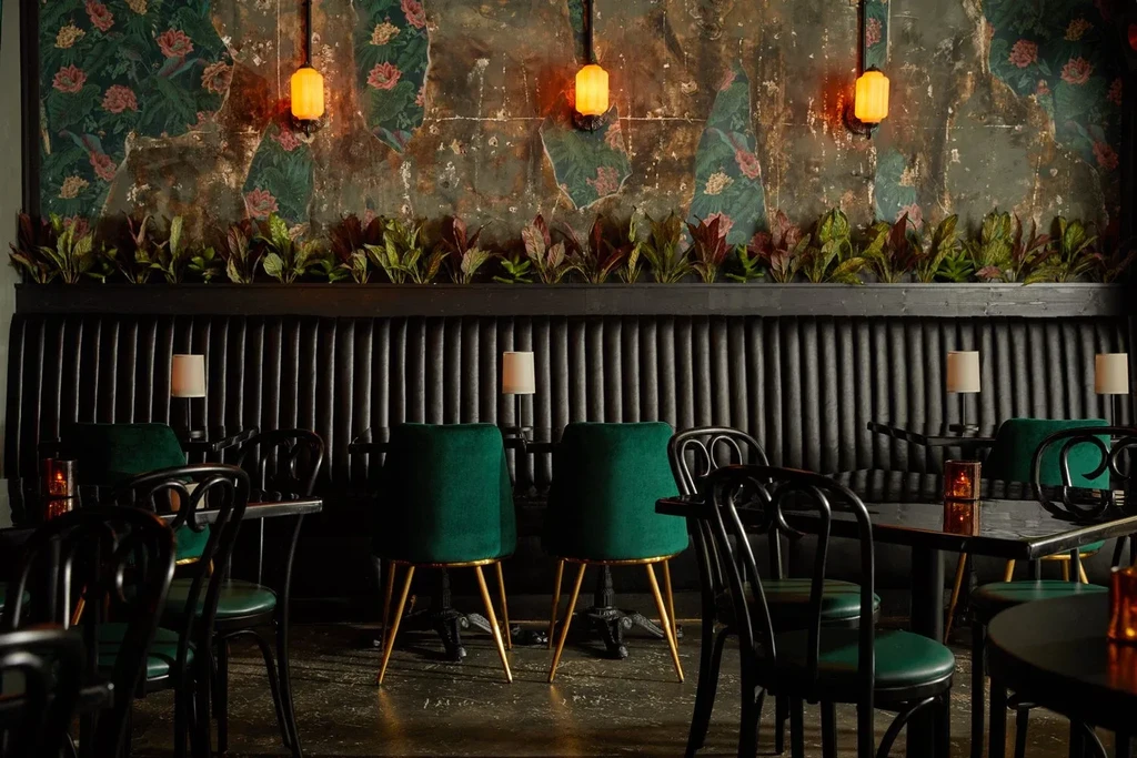 Moody restaurant interior with green chairs, banquette, and distressed floral wall.