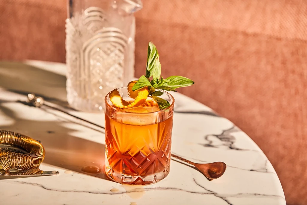 Amber cocktail with orange peel and basil, on marble with barware.