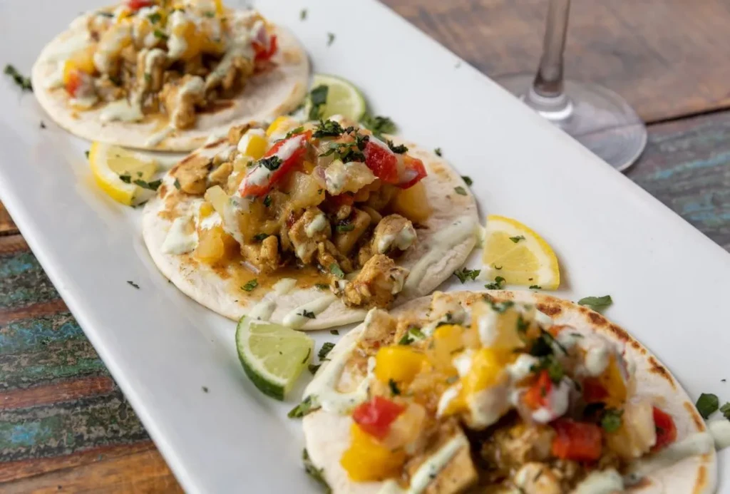 Three colorful tacos with seasoned protein, peppers, fruit, and lime.