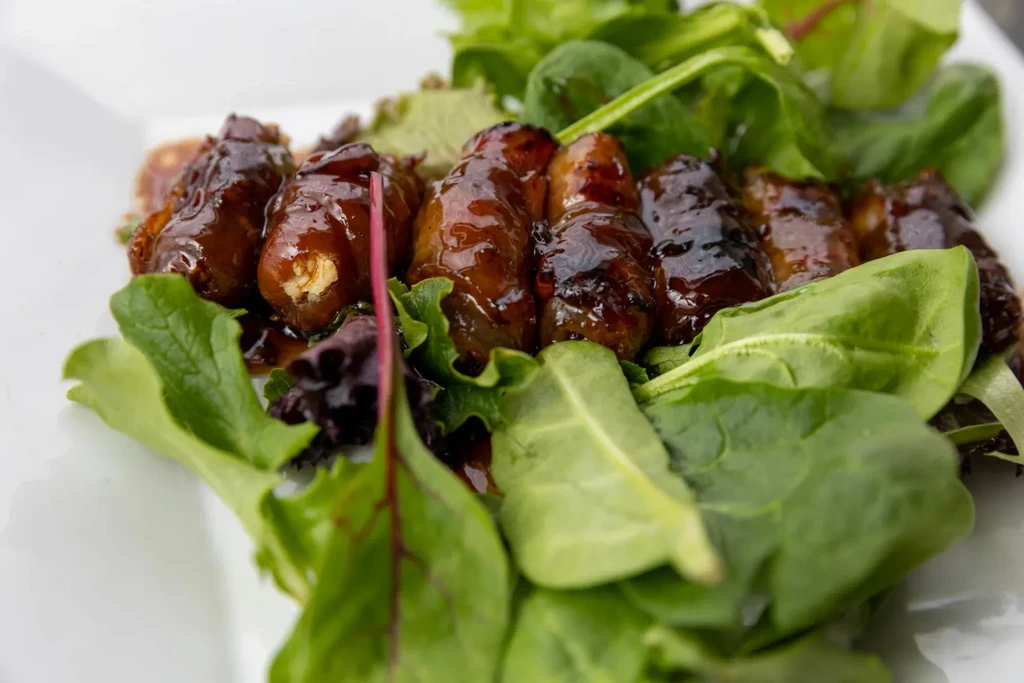 Darkly glazed bacon-wrapped dates on mixed greens, white plate.