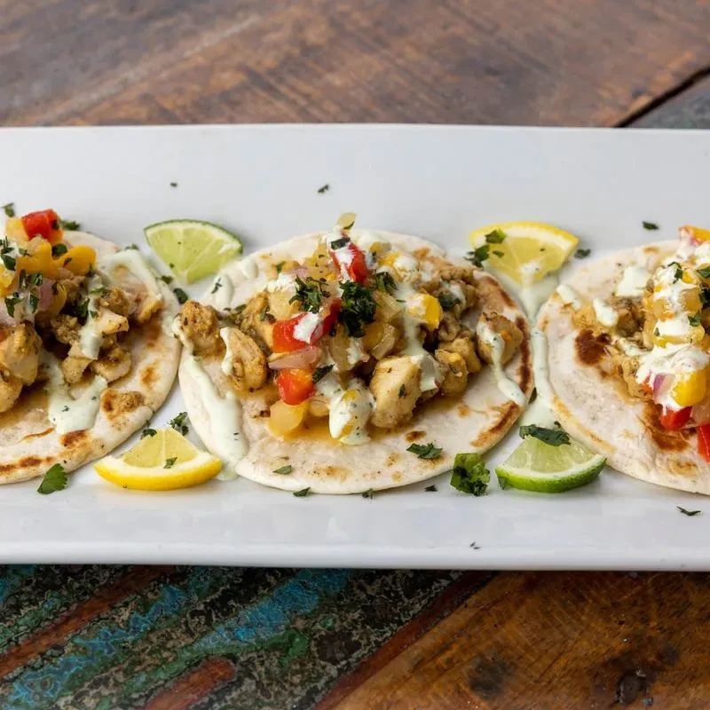 Three chicken tacos topped with peppers, sauce, herbs, and lime.