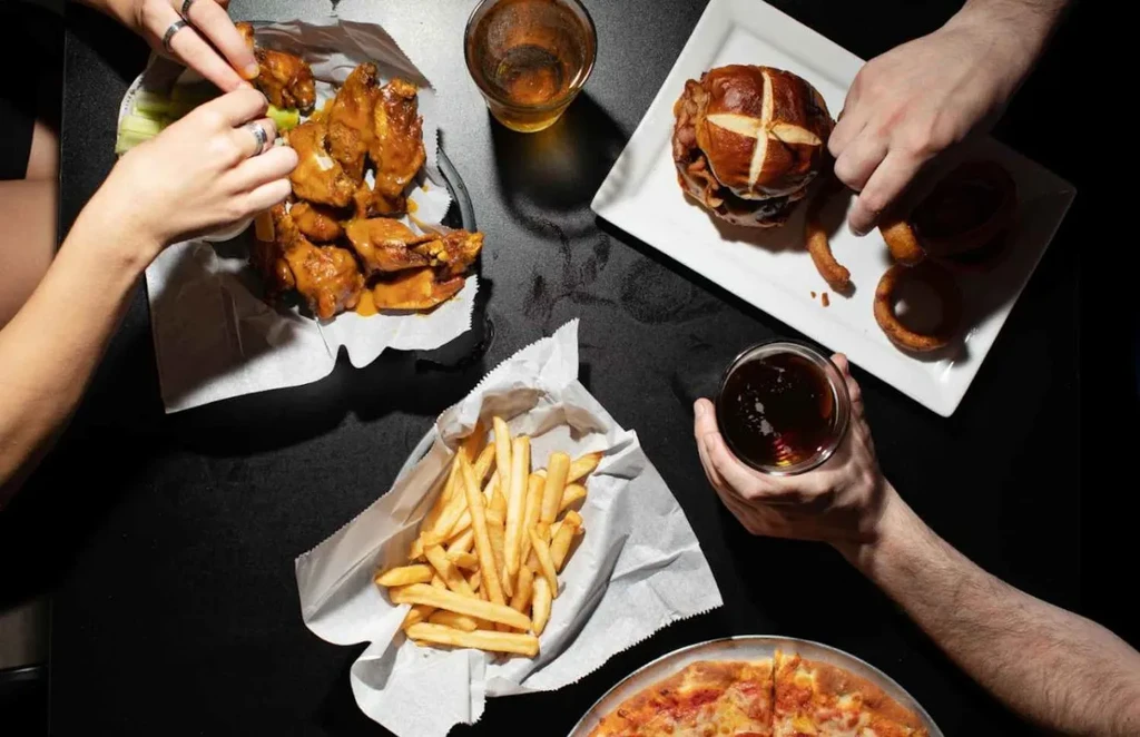 Hands reach for chicken wings, burger, fries, pizza, and drinks.