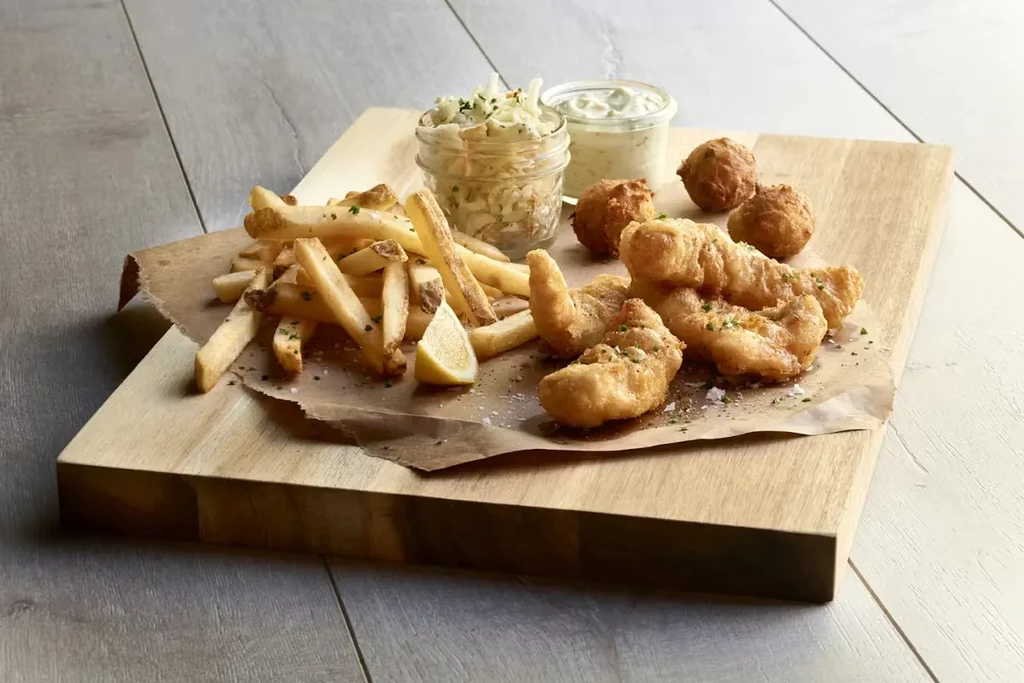 Fried fish, french fries, coleslaw, tartar sauce on wooden board.