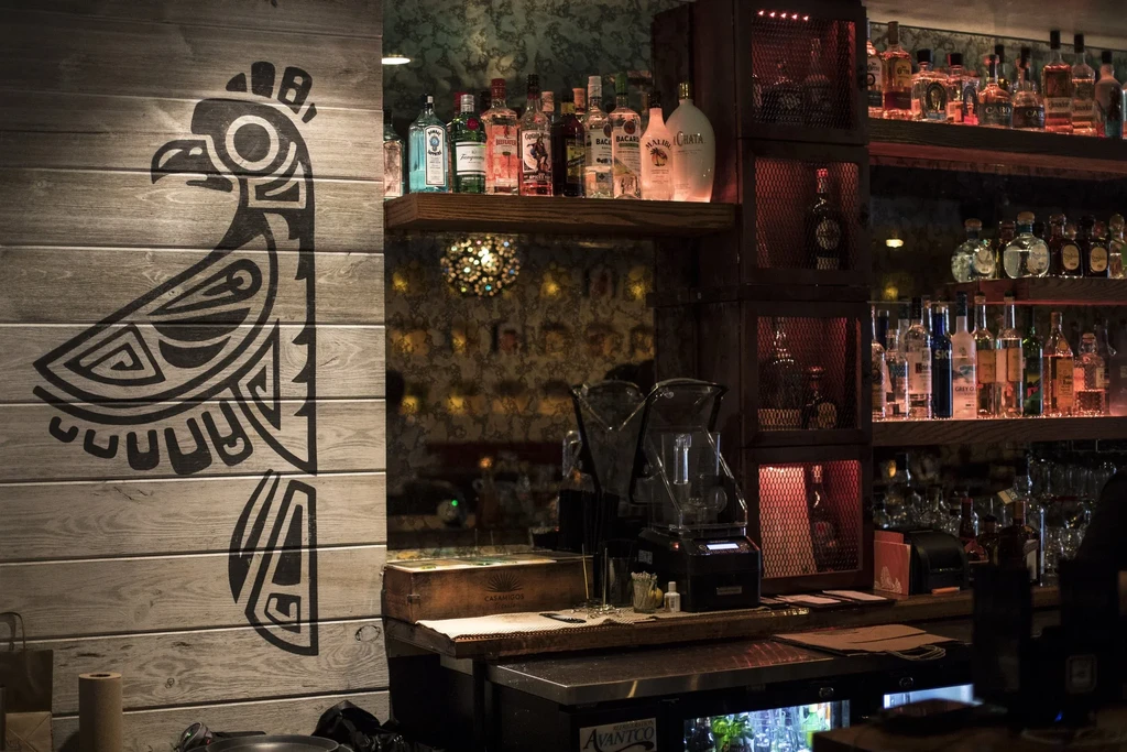 Warmly lit bar interior with a stylized bird drawing and liquor bottles.