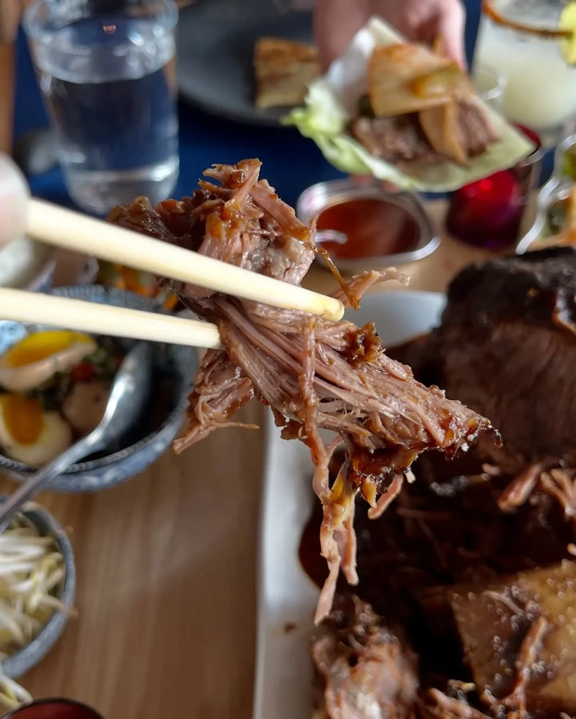Chopsticks lifting saucy pulled meat, blurred restaurant dishes behind.