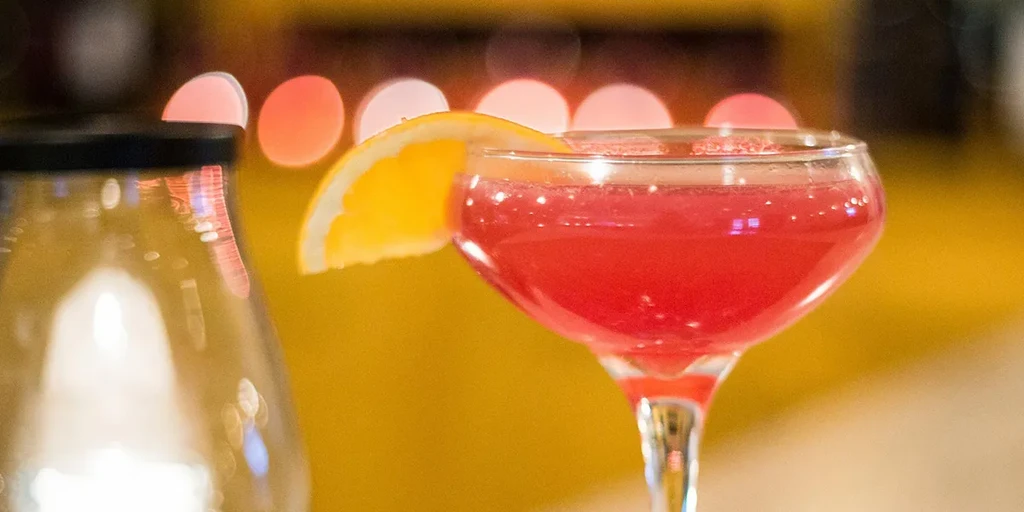 Fizzy red cocktail in coupe glass with orange, warm bokeh background.