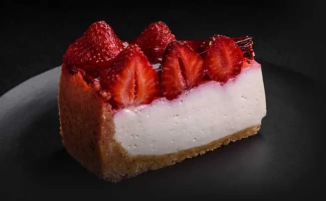 Slice of glazed strawberry cheesecake on dark background.