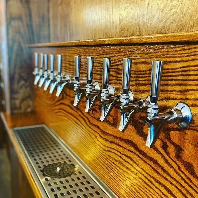 Multiple shiny chrome beer taps on a dark wooden bar with a drip tray.