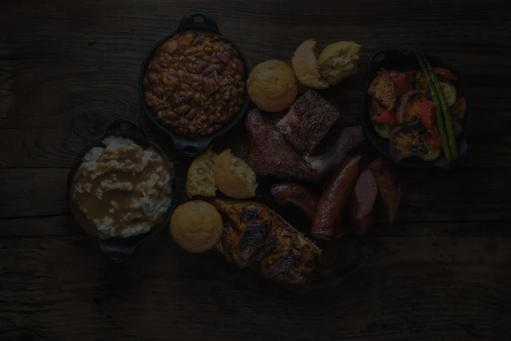 Rustic meal with meats, beans, mashed potatoes, cornbread on wood table.