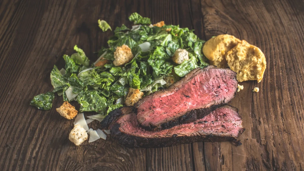Medium-rare steak, Caesar salad, and biscuits on a dark wooden board.