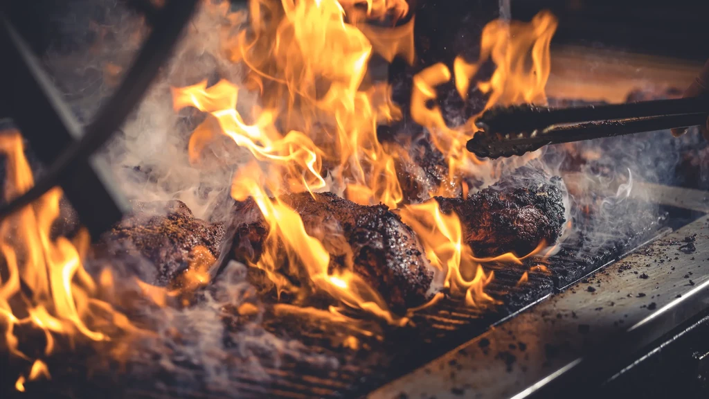 Close-up: Hand with tongs turns seared meat over intense grill flames.