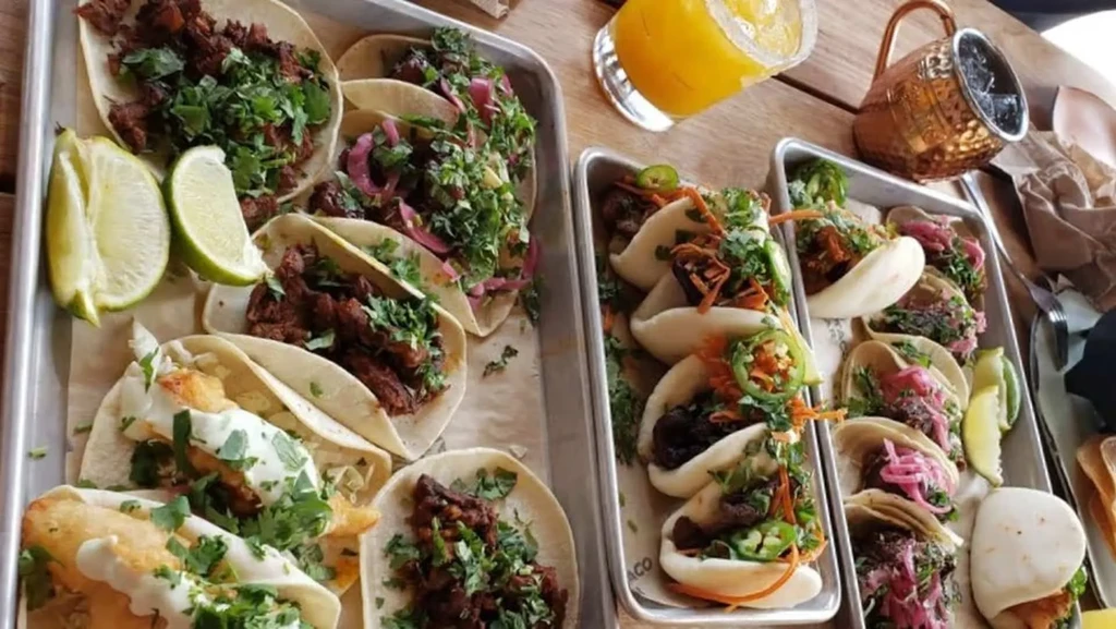 Overhead view of tacos, bao buns, and drinks on a wooden table.