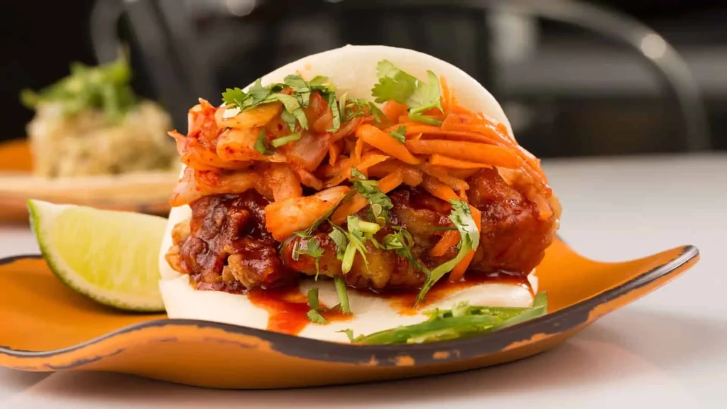 Fried chicken bao bun with kimchi and lime on a wavy ceramic plate.