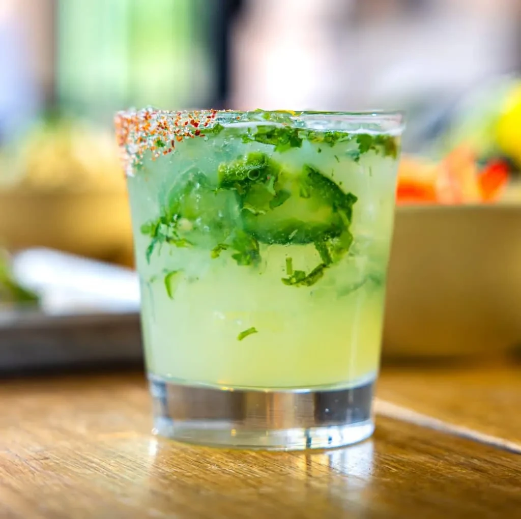 Green cocktail with ice, mint, and a chili-salted rim on wood.