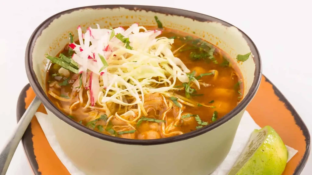 A bowl of red pozole soup, garnished with cabbage, radish, and lime.