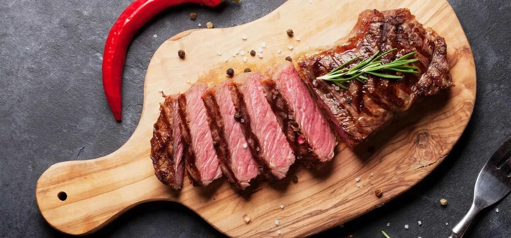 Partially sliced medium-rare steak on a wooden board with rosemary and seasoning.