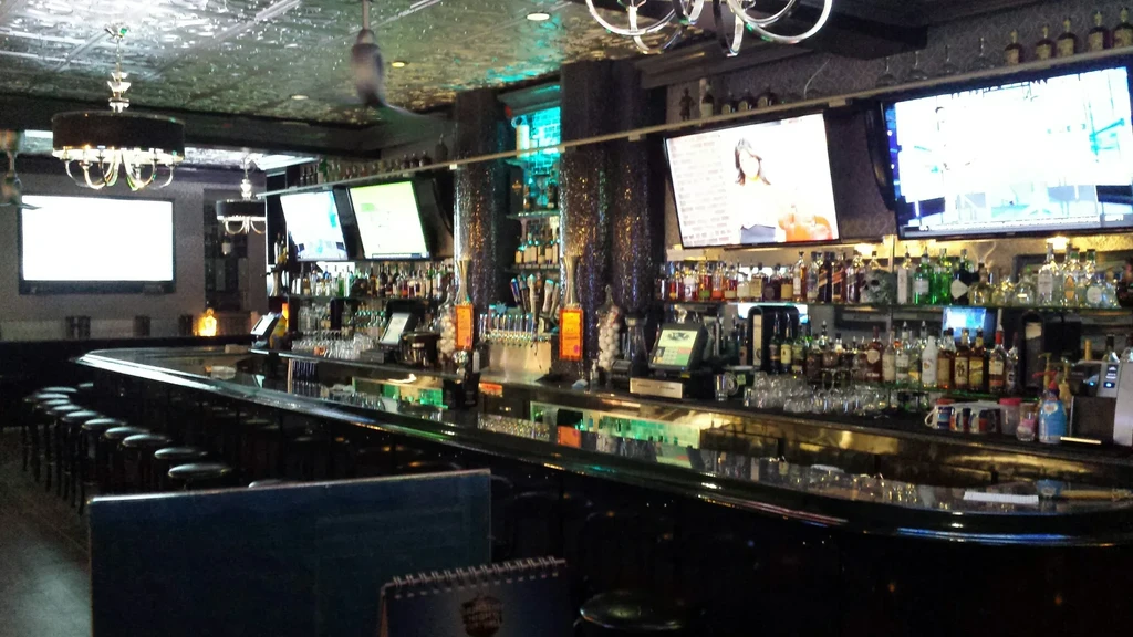 Dark, modern bar with curved counter, stools, liquor bottles, and TVs.