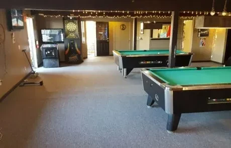 Two pool tables in an empty game room with arcade games and bar.