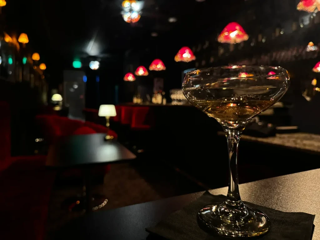 Close-up of amber cocktail on a dark bar, with colorful lamps.