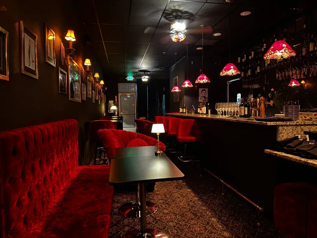 Elegant bar interior with red velvet banquette, dark bar, and ornate lights.