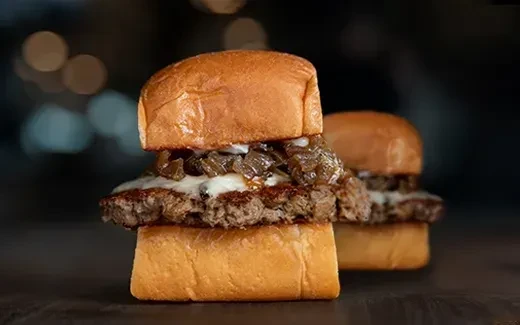 Two beef sliders with melted cheese and caramelized onions on a dark table.