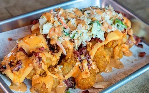 Tater tots, cheese sauce, bacon, and coleslaw in a metal tray.